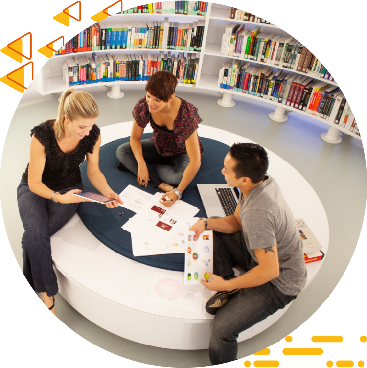 Aerial photo of three students collaborating on design work while seated on a large ottoman, 书籍是背景.